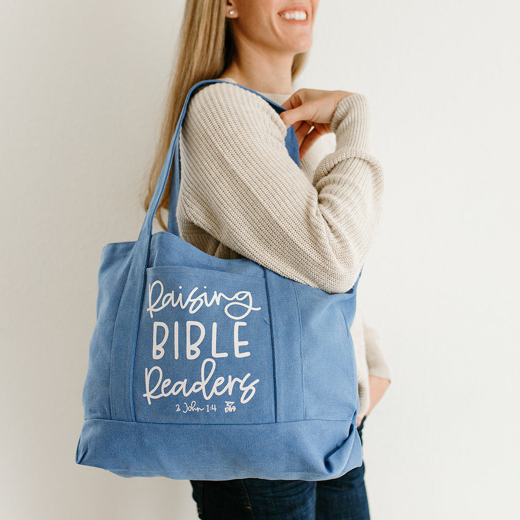 Woman holding a blue tote bag that says Raising Bible Readers, against a plain background